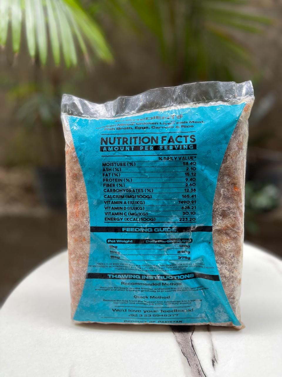 Backside of MunchBowl - Canine Naturals Pakistan. A bag of wet cat food with a prominent blue nutrition facts label, standing on a white surface with blurred green foliage in the background.