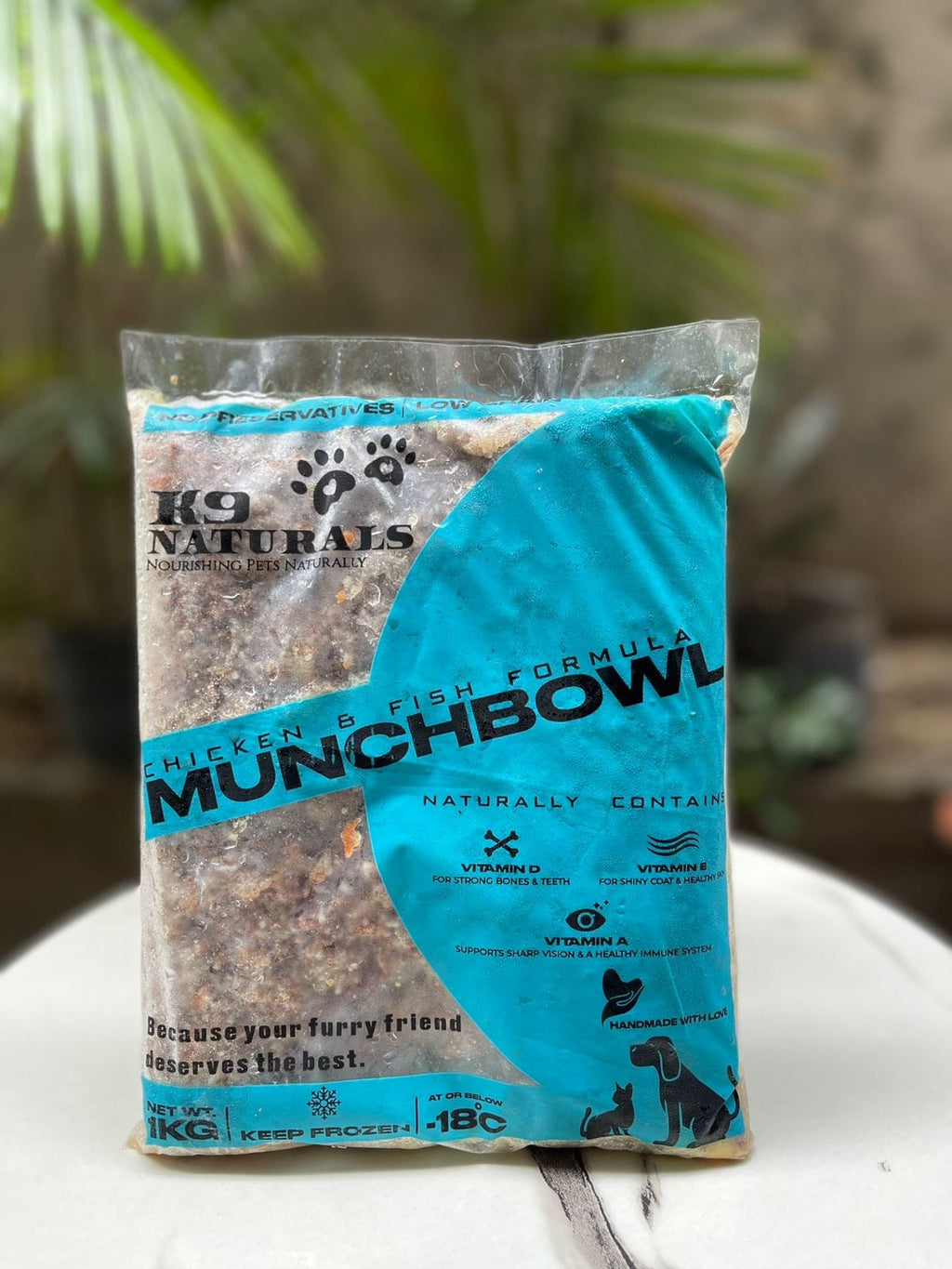 A 1kg bag of K9 Naturals Munchbowl Chicken & Fish Formula wet dog food, displayed on a white surface with a blurred background of green foliage.